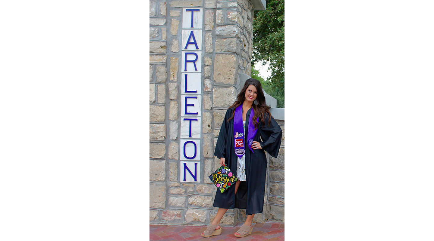 Shelbi Tidwell wearing her purple graduation gown, holding her graduation cap next to a Tarleton State University sign. 